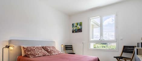Un dormitorio con cama, una silla, una mesa, una lámpara, un radiador y una ventana con vistas.