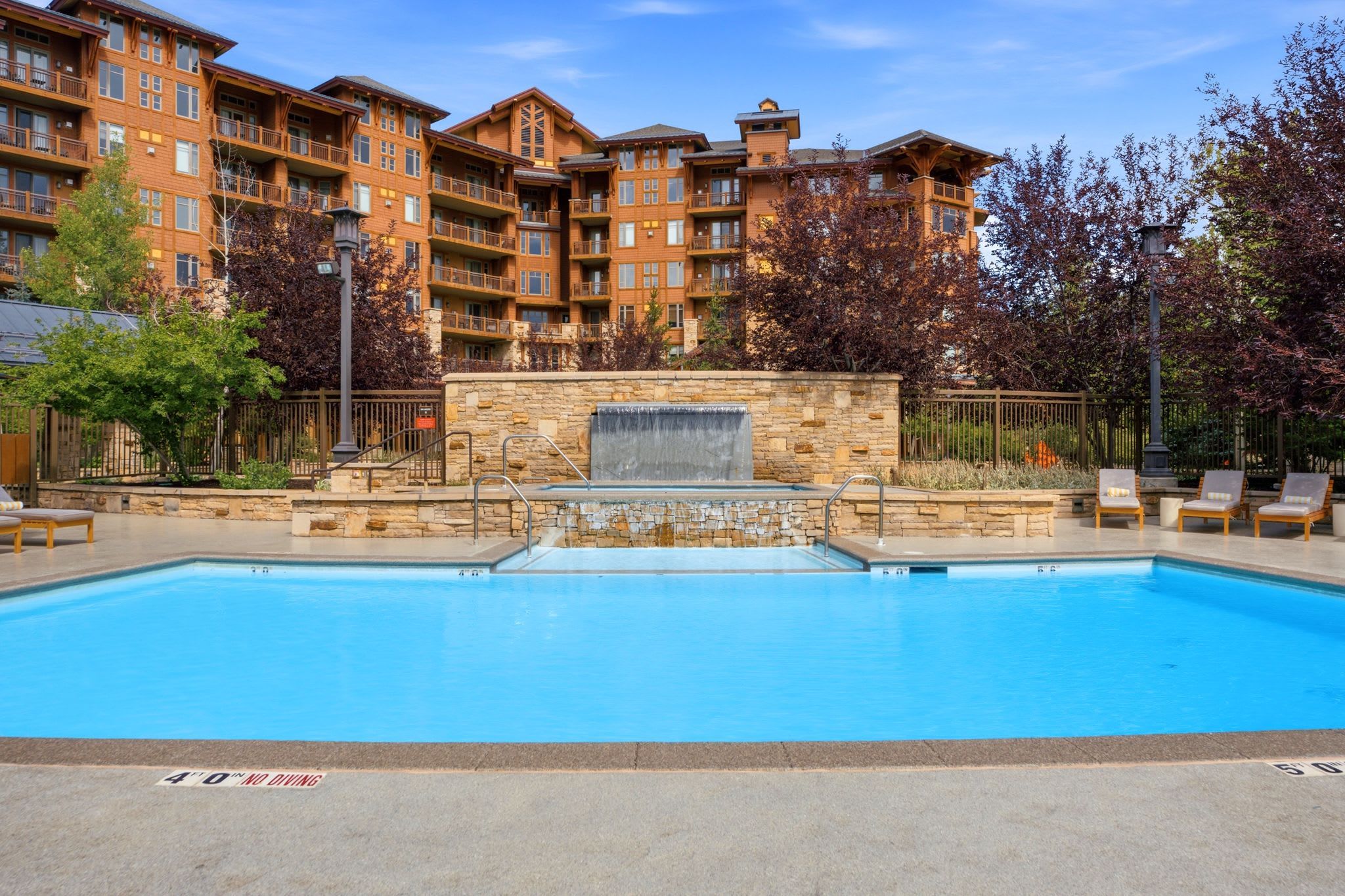 A swimming pool with a stone waterfall feature, surrounded by a stone wall and a wooden fence.