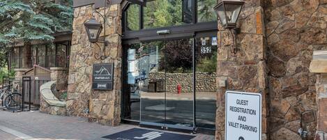 A building entrance with a sign for guest registration and valet parking, a stone facade, and a doormat with a logo.