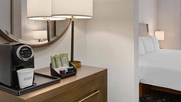 A hotel room with a bed, a desk with a coffee machine, a lamp, and a window with curtains.