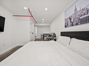 Ein modernes Hotelzimmer mit einem großen Bett, einem grauen Sessel, einem kleinen Tisch und einem Stadtbild an der Wand.