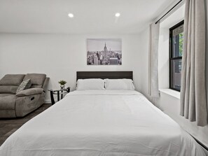 Un dormitorio con cama, un sillón reclinable, una mesita con una planta y una ventana con cortinas.