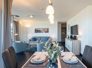 A modern living room with a sofa, dining table set for two, and a kitchen area with a TV. - Holiday Apartment With 1 Bedroom (Heiligenhafen)