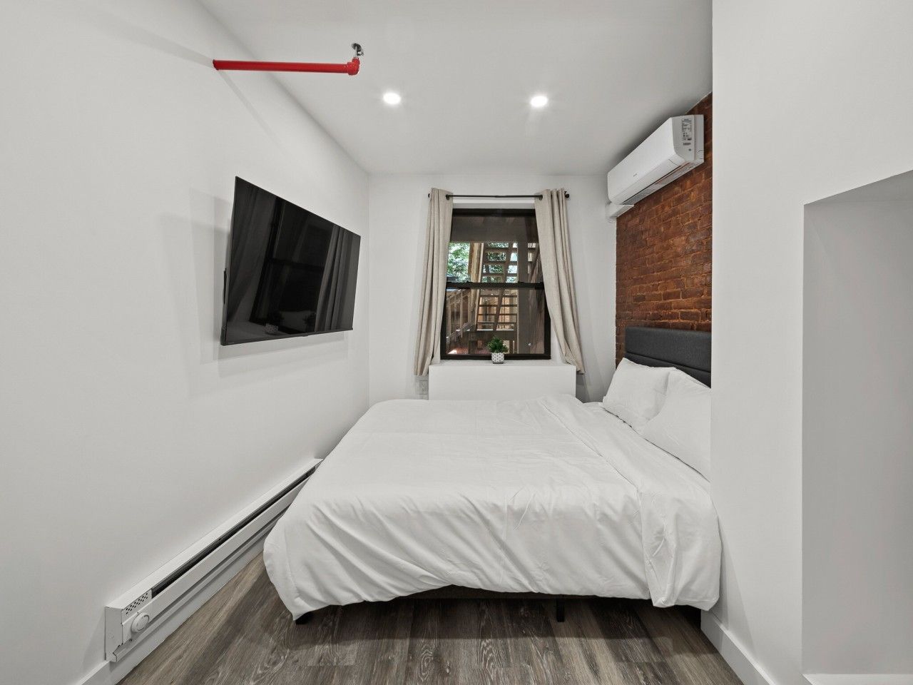 Un dormitorio con cama, ventana con cortinas, televisor montado y unidad de aire acondicionado.
