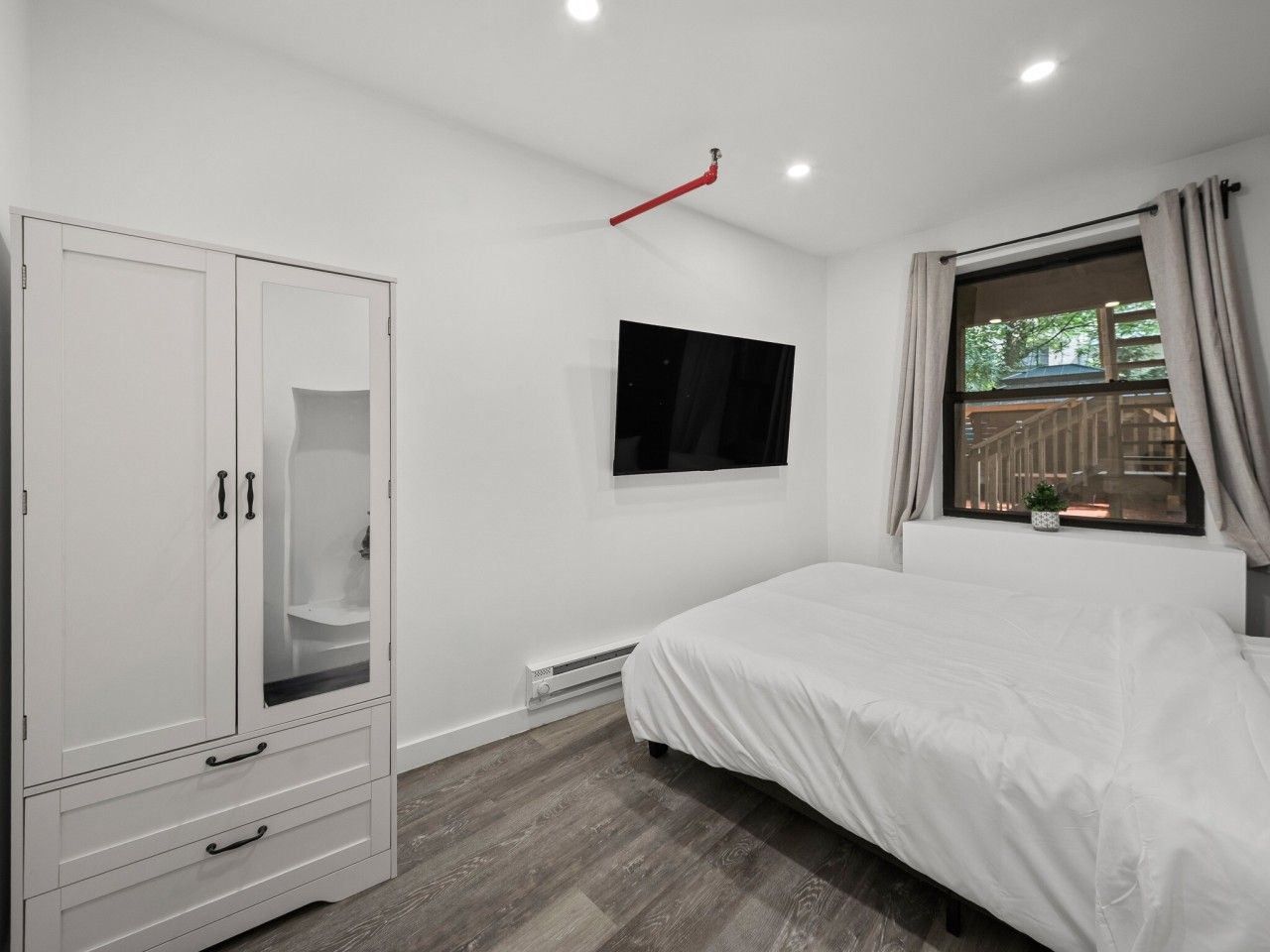 Ein Schlafzimmer mit Bett, Kleiderschrank, Fernseher, einem Fenster mit Vorhängen und einer Heizung.