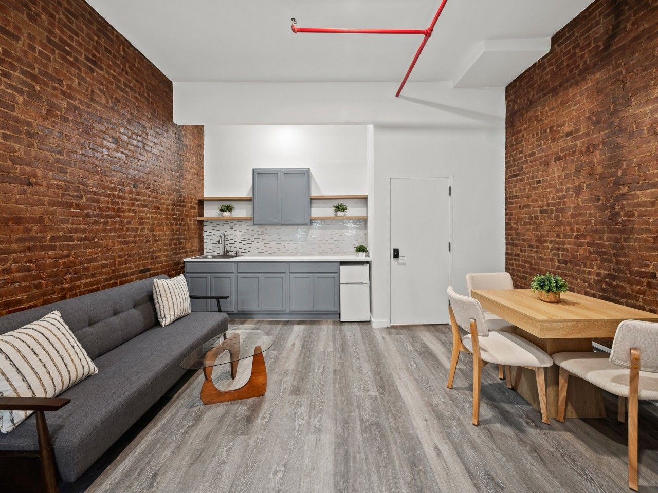 Eine moderne Küche mit grauen Schränken und einer weißen Arbeitsplatte, ein Essbereich mit einem Holztisch und -stühlen sowie ein graues Sofa mit gestreiften Kissen.