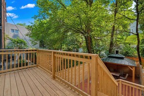 A wooden deck with a railing, surrounded by trees and a small building with a green roof.