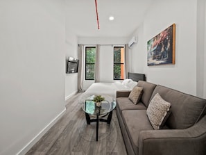 A modern hotel room with a bed, a sofa, a glass coffee table, and a painting on the wall. - Spacious King Room with Full-Size Sleeper Sofa (New York)