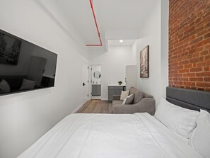 A modern bedroom with a bed, a grey sofa, a small kitchenette, and a brick wall.