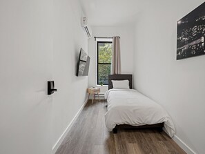 A bedroom with a bed, a window with curtains, a small table, and a wall-mounted air conditioner.