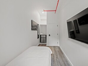 Ein schmales Hotelzimmer mit einem Bett, einem kleinen Tisch, einem Spiegel und einem Fenster.