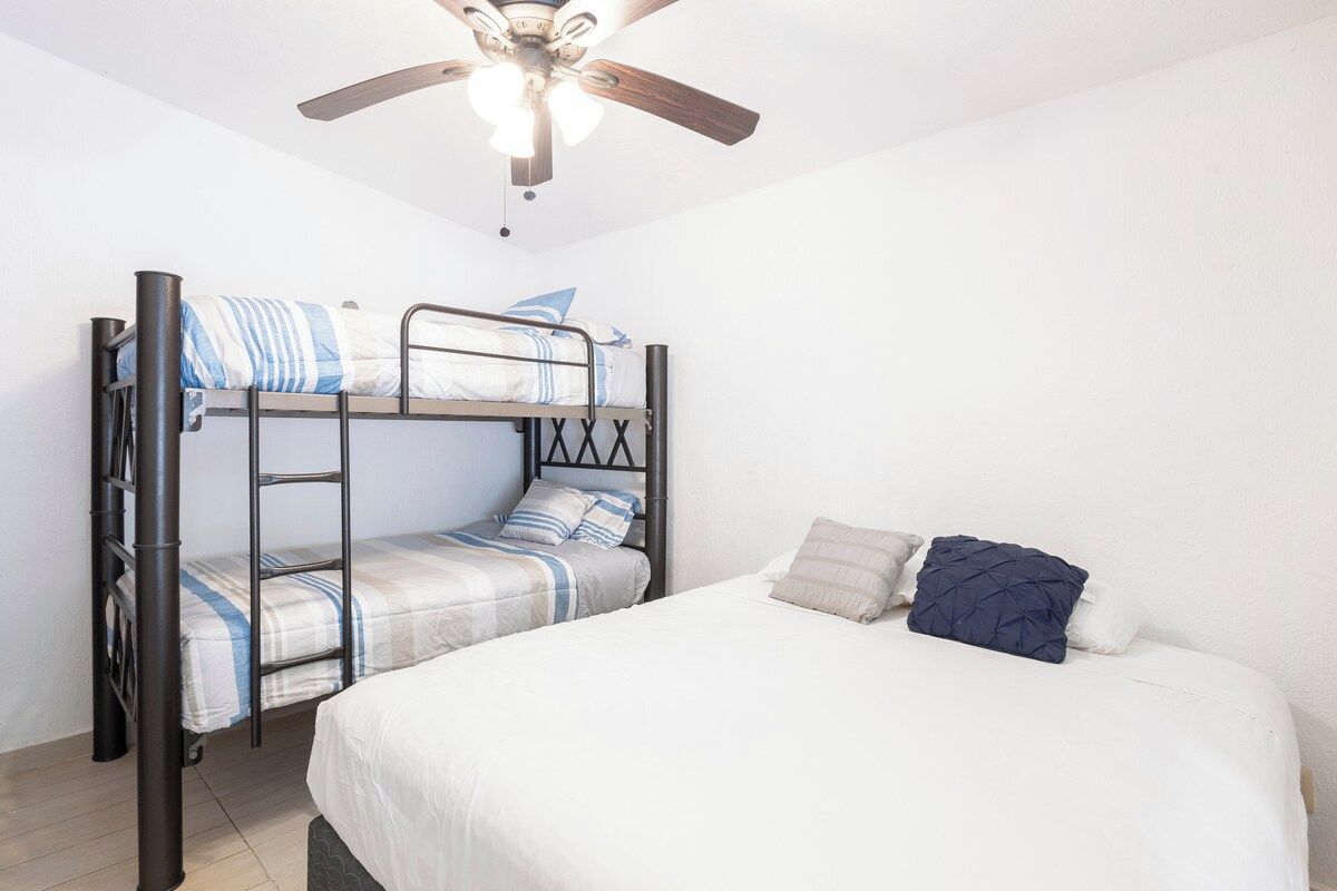 Una cama litera con escalera, una cama individual y un ventilador de techo en una habitación con paredes blancas.