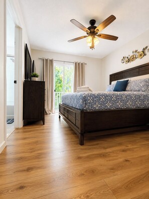 Une chambre avec un plancher en bois, un lit avec une literie à motifs, un ventilateur de plafond et une fenêtre avec des rideaux.