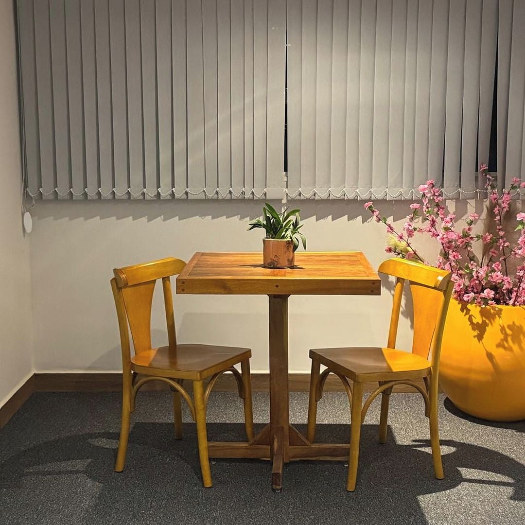 Una habitación con una mesa de madera y dos sillas, una planta en maceta sobre la mesa, y una maceta amarilla grande con flores rosadas.