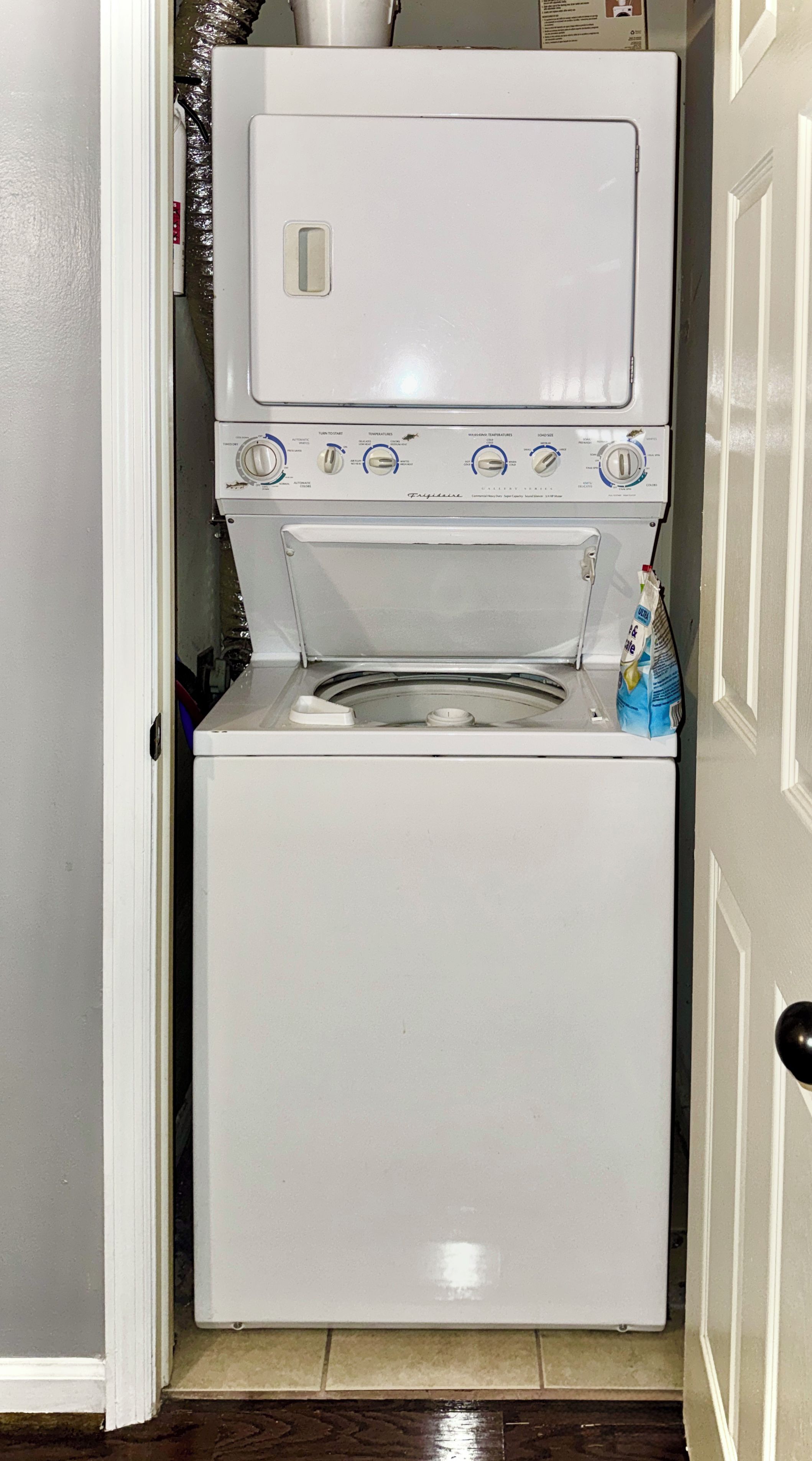 Een compacte, witte wasmachine met voorlaadopening en bedieningspaneel, in een smalle gang met een deur aan de rechterkant.