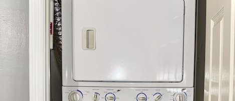 A compact, white front-loading washing machine with a control panel, located in a narrow hallway with a door on the right.