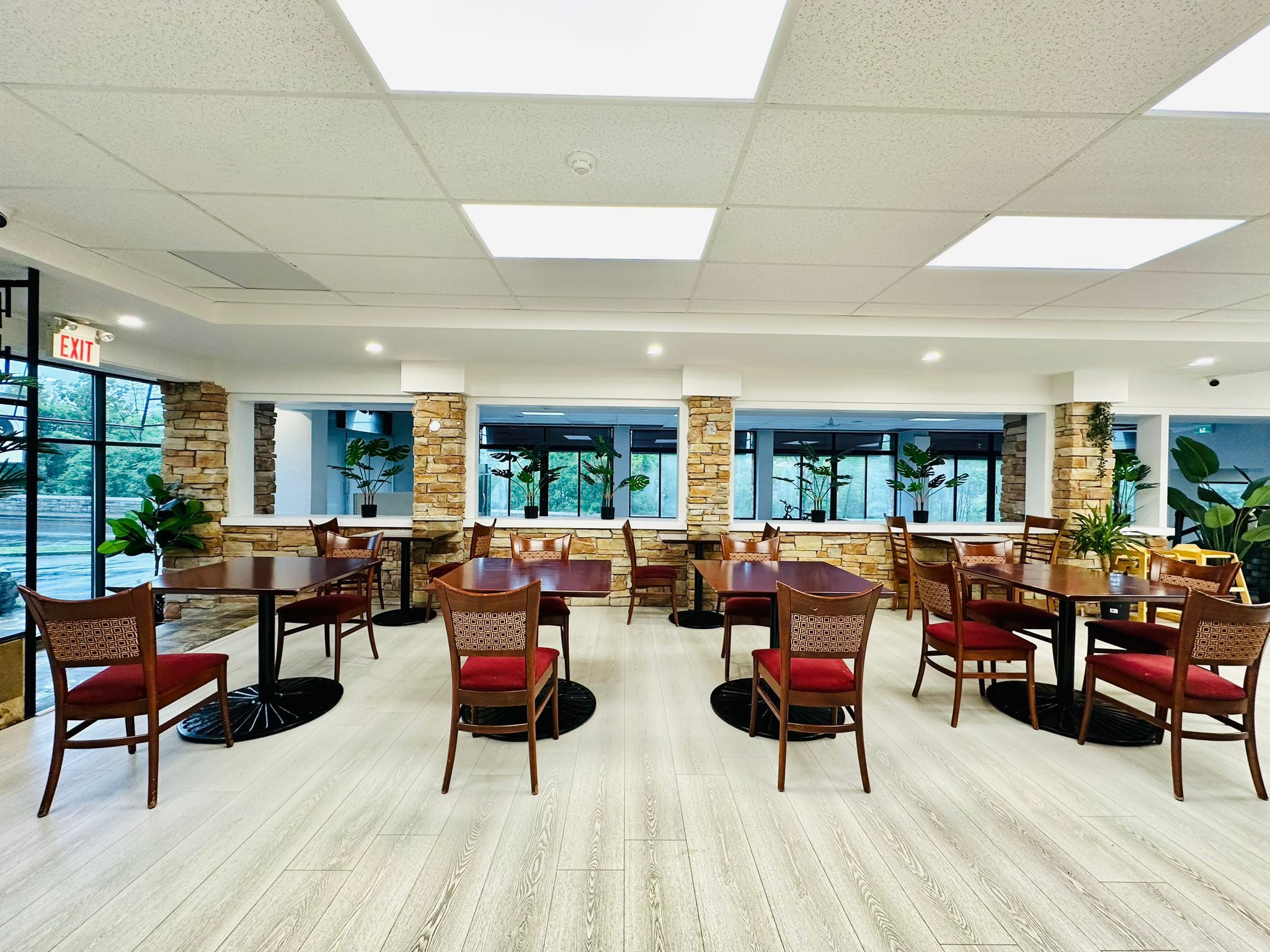 A spacious dining area with multiple tables and chairs, large windows, and potted plants.
