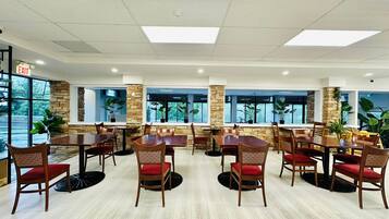 A spacious dining area with multiple tables and chairs, large windows, and potted plants.