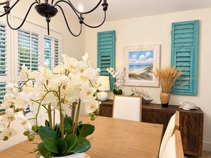 A dining area with a wooden table, white orchids, a painting of a beach, and teal shutters on the walls.