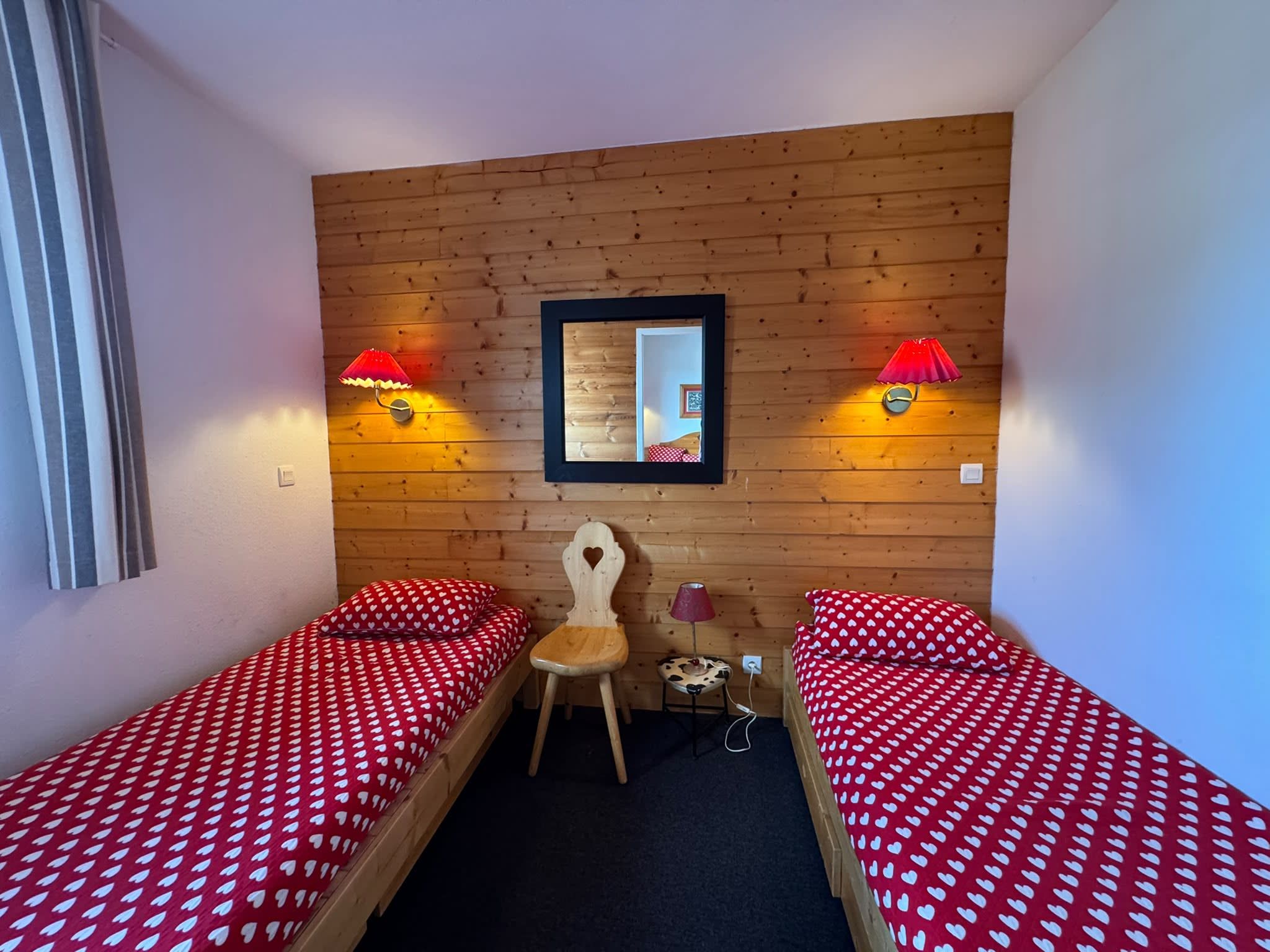 Une petite pièce avec deux lits simples, une tête de lit en bois, une chaise, une petite table et deux lampes fixées au mur.