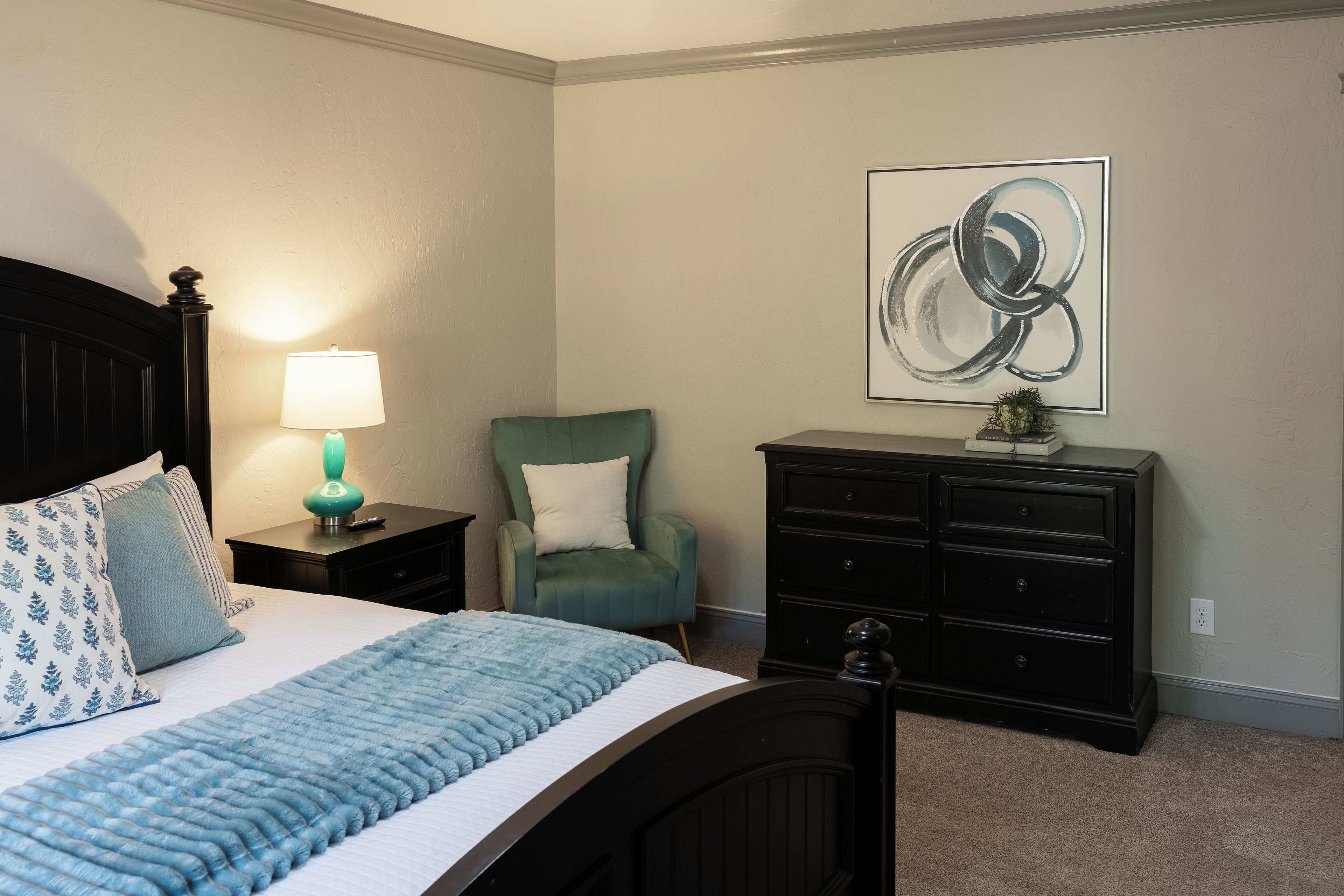A bedroom with a bed, a nightstand, a lamp, a chair, a dresser, and a wall art piece.