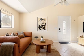 Una sala de estar con un sofá, una mesa redonda de madera, una puerta con paneles de vidrio decorativos y una obra de arte enmarcada en la pared.