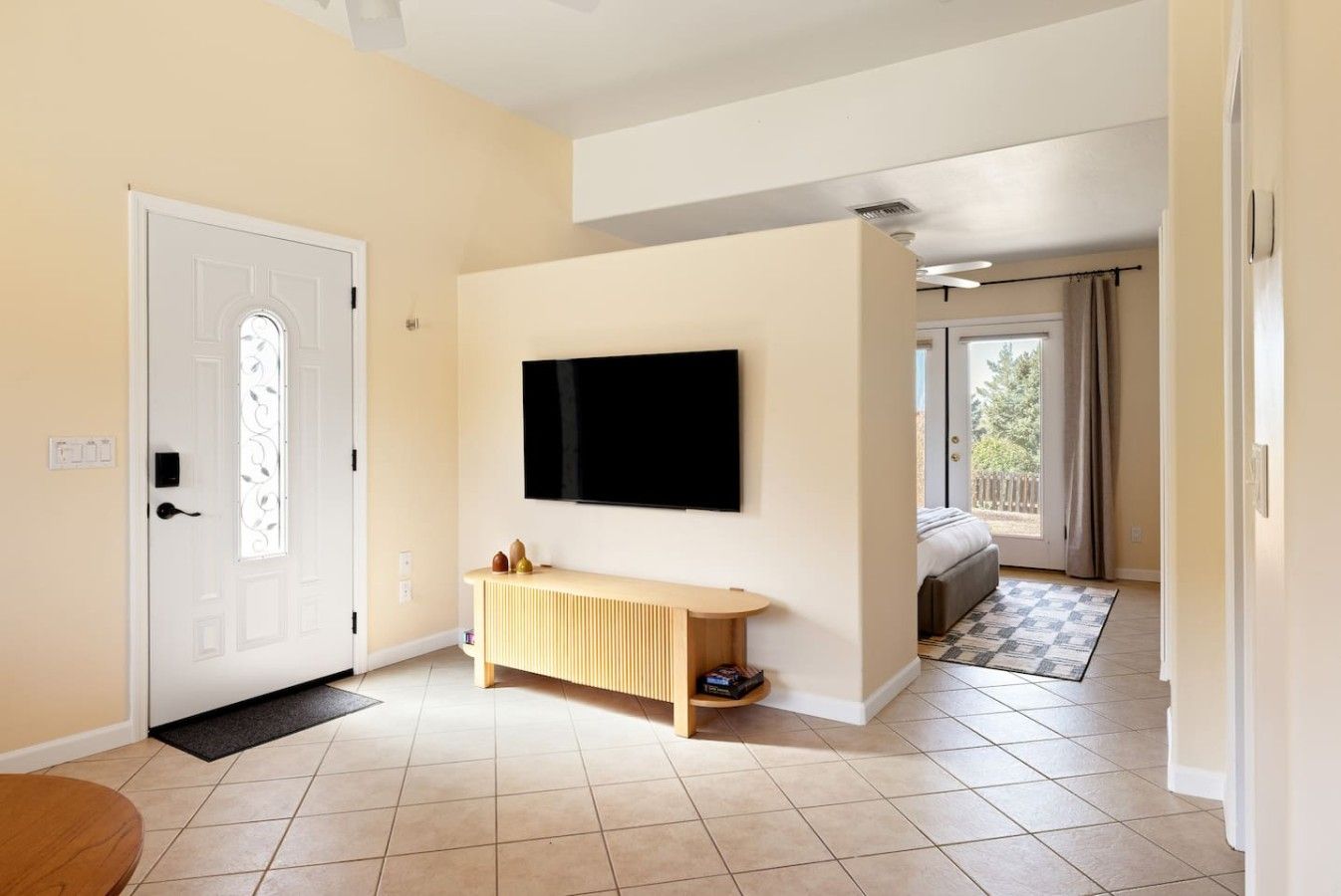 A modern living room with a flat-screen TV mounted on the wall, a wooden console table, and a large window offering a view of the outdoors.