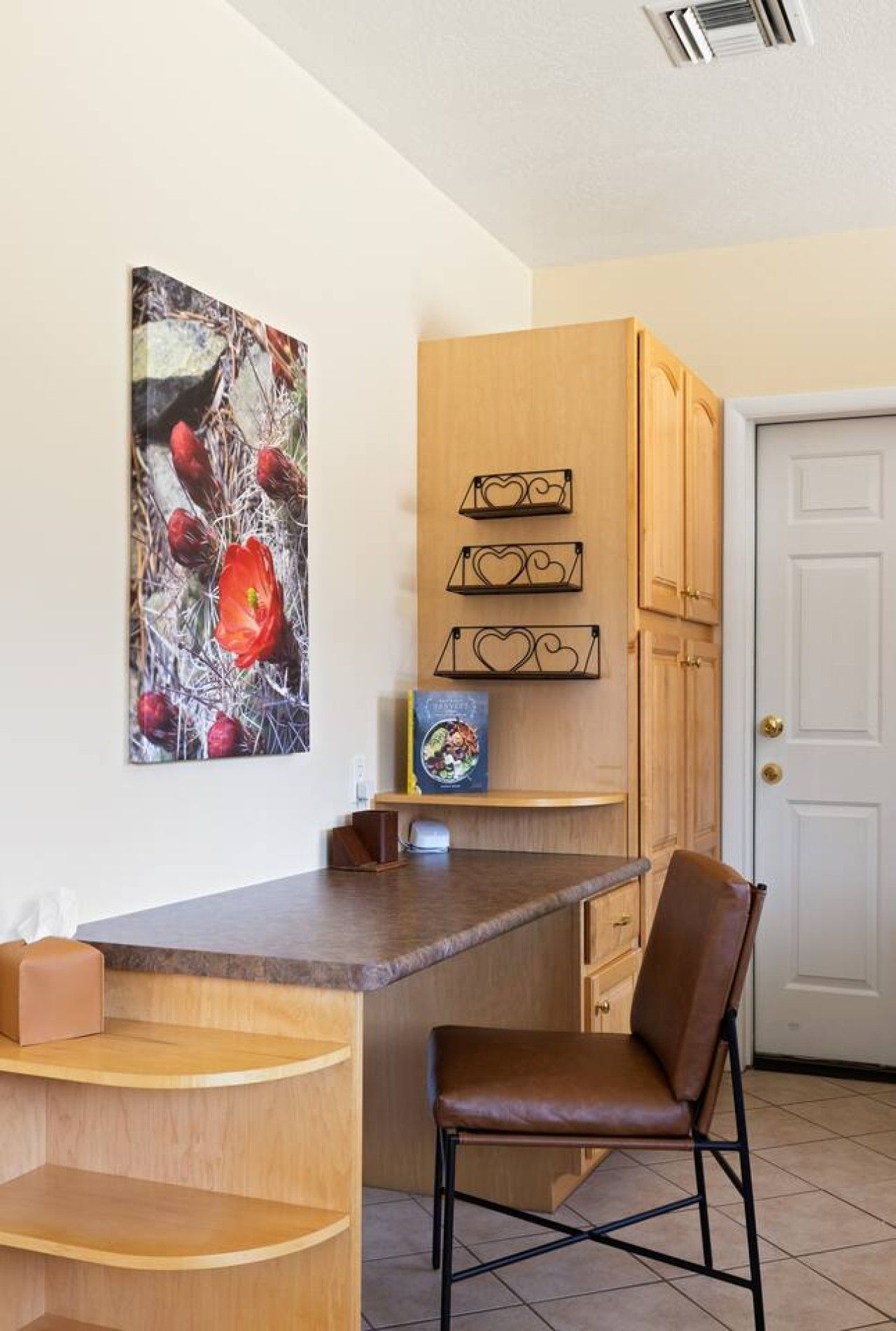 A kitchen with wooden cabinets, a white door, a brown chair, and a painting of red flowers.