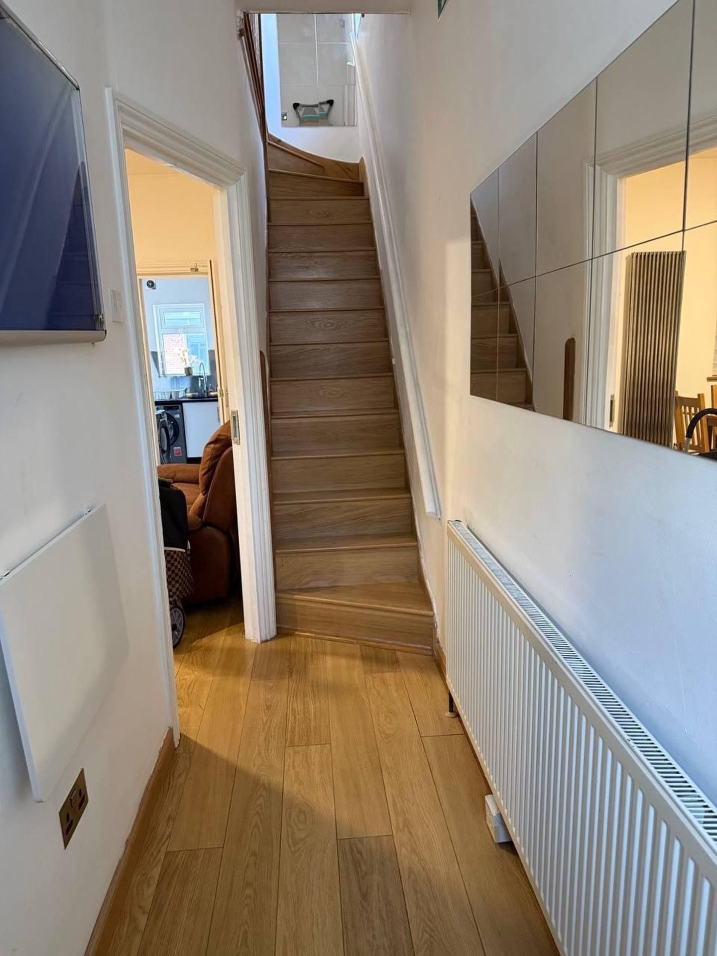 A hallway with a wooden staircase, a radiator, and a door leading to a room with a bed and a chair.