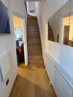 Un pasillo con una escalera de madera, un radiador y una puerta que da a una habitación con cama y sillón.