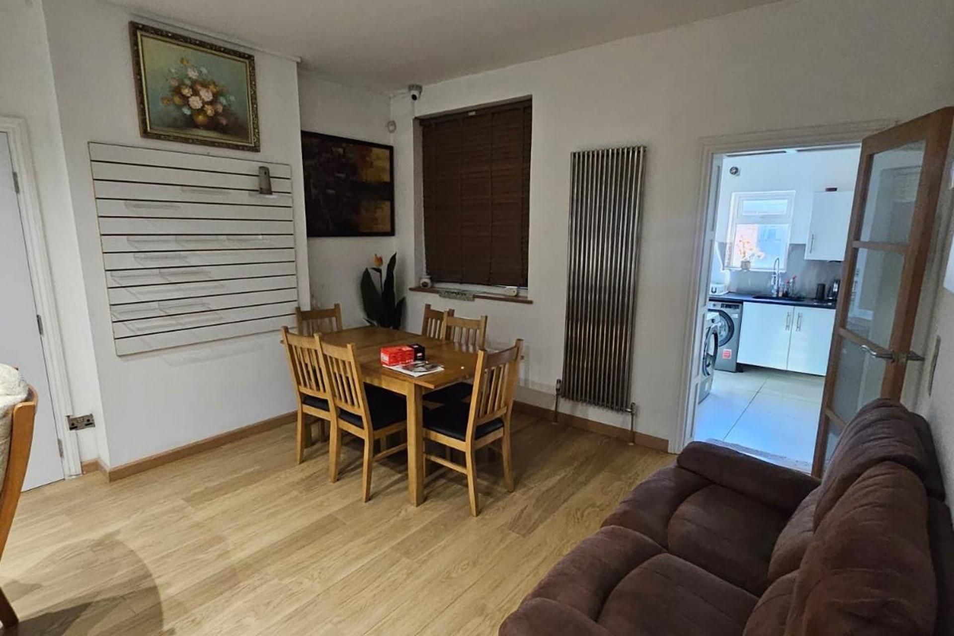 Un área de comedor con una mesa y sillas de madera, un sofá marrón y una cocina visible a través de una puerta abierta.
