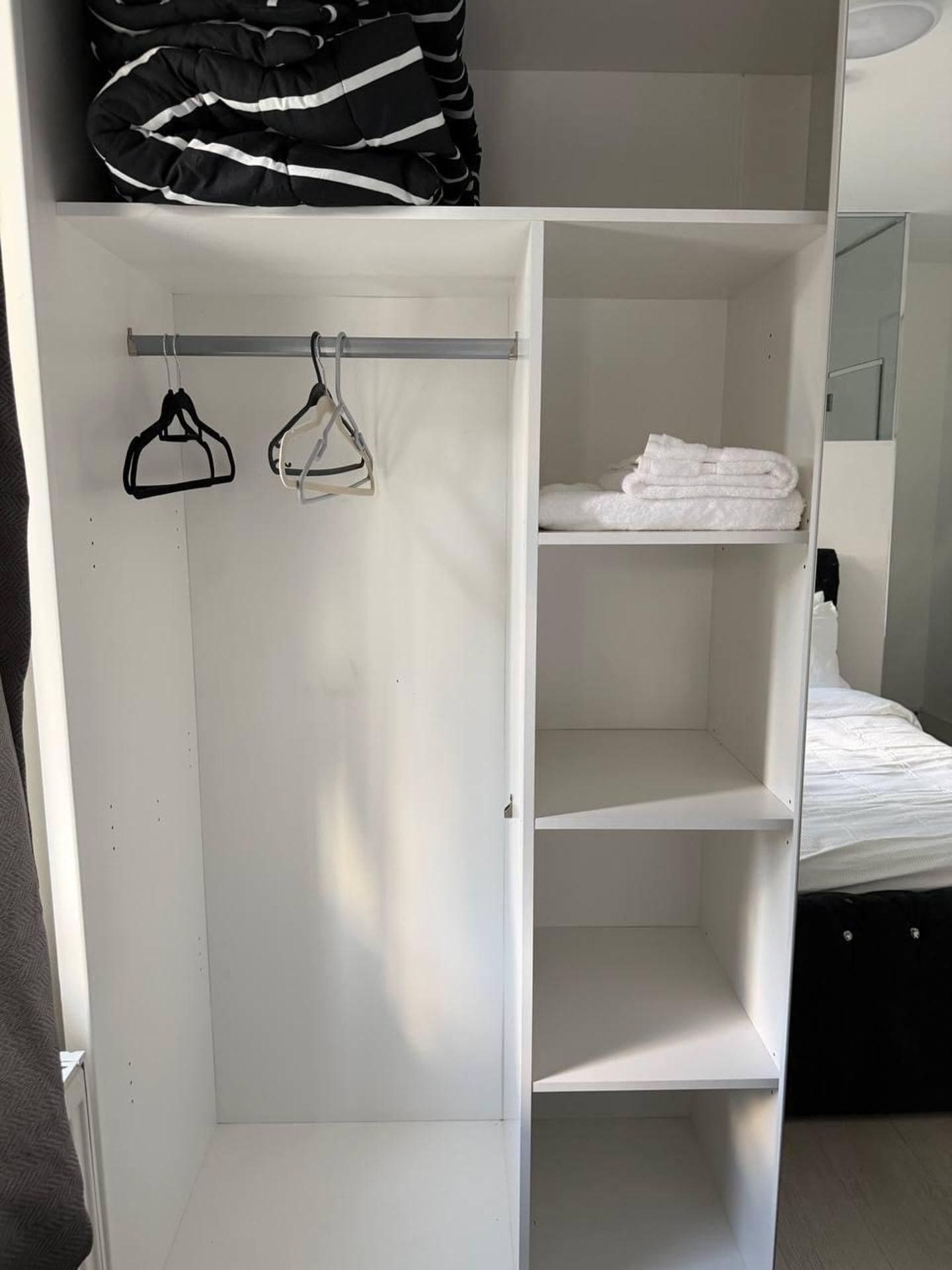 A white wardrobe with hanging space and shelves, a bed with white bedding, and a mirror.