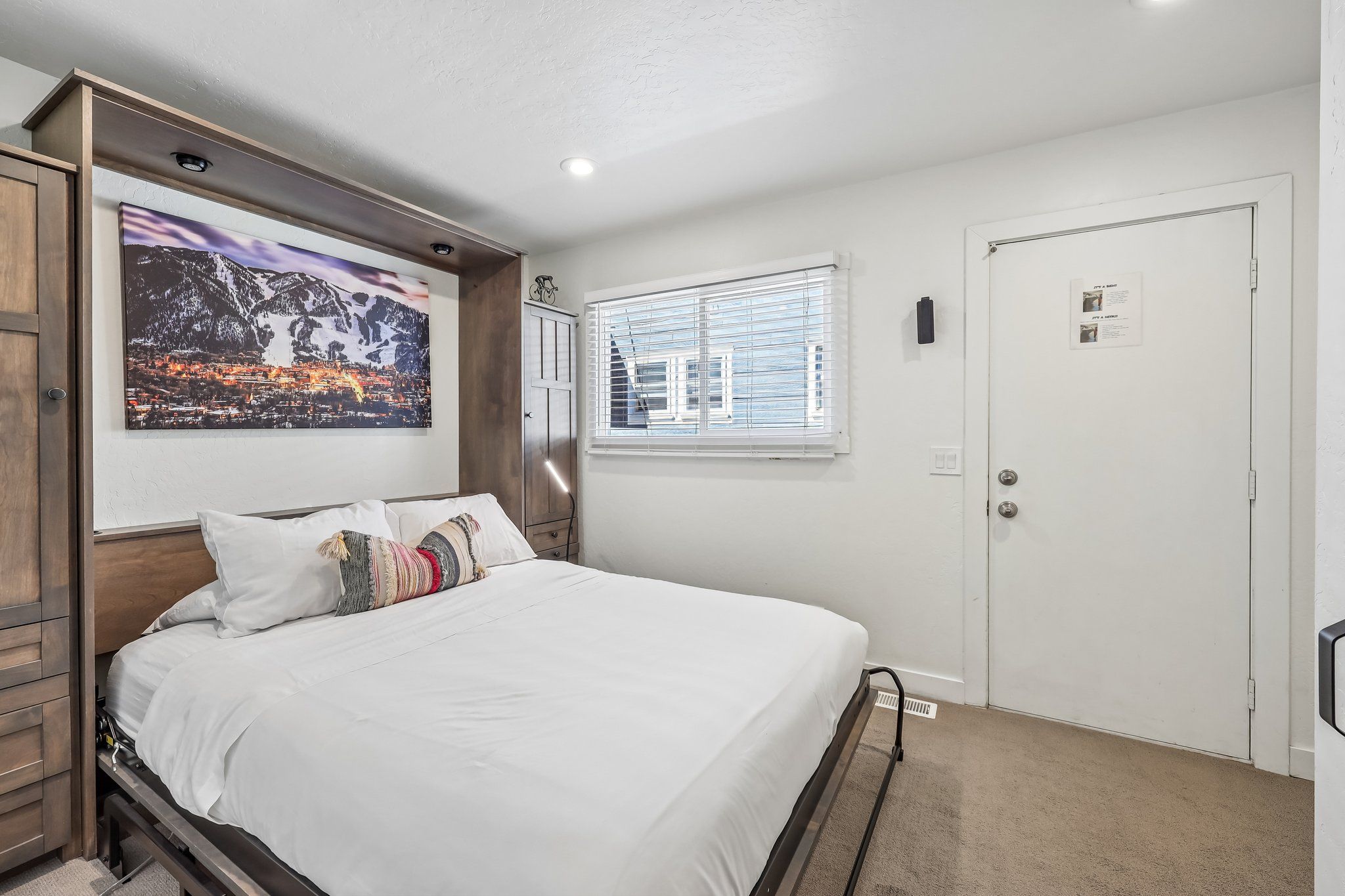 A bedroom with a bed, a window with blinds, a door, and a wall-mounted picture.