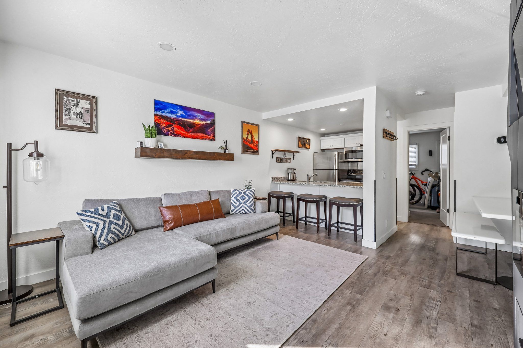 A modern living room with a grey sectional sofa, a coffee table, and a kitchen area with a fridge, microwave, and bar stools.