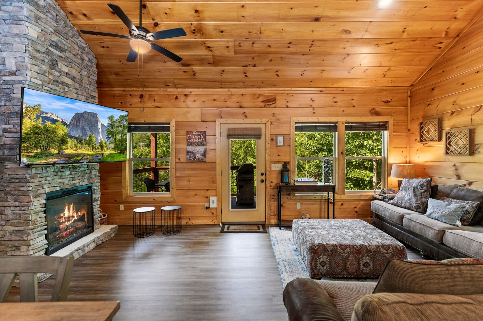 Ein Interieur einer Hütte mit Kamin aus Stein, Deckenventilator, einer Couch und einem Fernseher, der eine Berglandschaft zeigt.