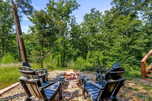 A circular fire pit with four chairs around it, set on a bed of pebbles, surrounded by a forested area. - The Sporting Place - Outdoor Hot Tub, Firepit, Game Room + Close to Pigeon Forge (Sevierville)