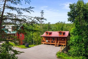 A cabin with a red roof, surrounded by dense forest, and a paved driveway leading to the entrance. - The Sporting Place - Outdoor Hot Tub, Firepit, Game Room + Close to Pigeon Forge (Sevierville)