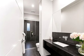 Une salle de bain moderne avec une douche carrelée de couleur sombre, des toilettes blanches et un plan de toilette noir agrémenté d’un lavabo blanc et d’un vase de fleurs.