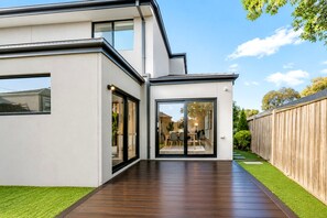 Une maison moderne à deux étages, dotée d’une terrasse en bois, de grandes portes vitrées donnant sur un coin repas et d’un jardin clôturé.
