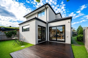 Une maison moderne à deux étages, dotée d’une terrasse, d’un coin repas visible à travers des portes vitrées et d’une pelouse bien entretenue.
