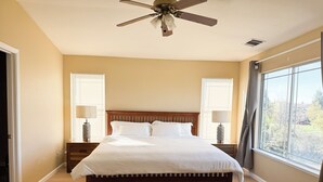 Una camera da letto con un letto di legno, due comodini con lampade, una ventola a soffitto e un'ampia finestra con persiane.