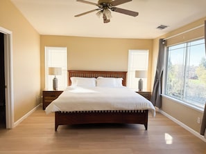 Un dormitorio con una cama de madera, dos mesitas de noche con lámparas, un ventilador de techo y un ventanal con persianas.