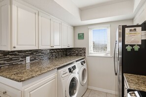 A kitchen with white cabinets, a granite countertop, a built-in washer and dryer, a black refrigerator, and a window with blinds. - Gulf Dunes 1 | 2 Bed-2.5 Bath | Gulf Front | Pet friendly! | Sleeps 5 (Boca Grande)