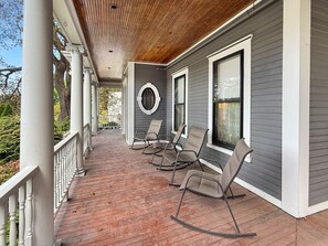 A covered porch with a wooden ceiling, grey siding, and a circular window. There are four rocking chairs and a white railing.