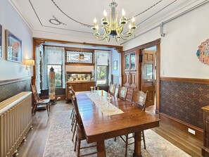 A formal dining room with a long wooden table, chairs, a chandelier, and a view through the windows.