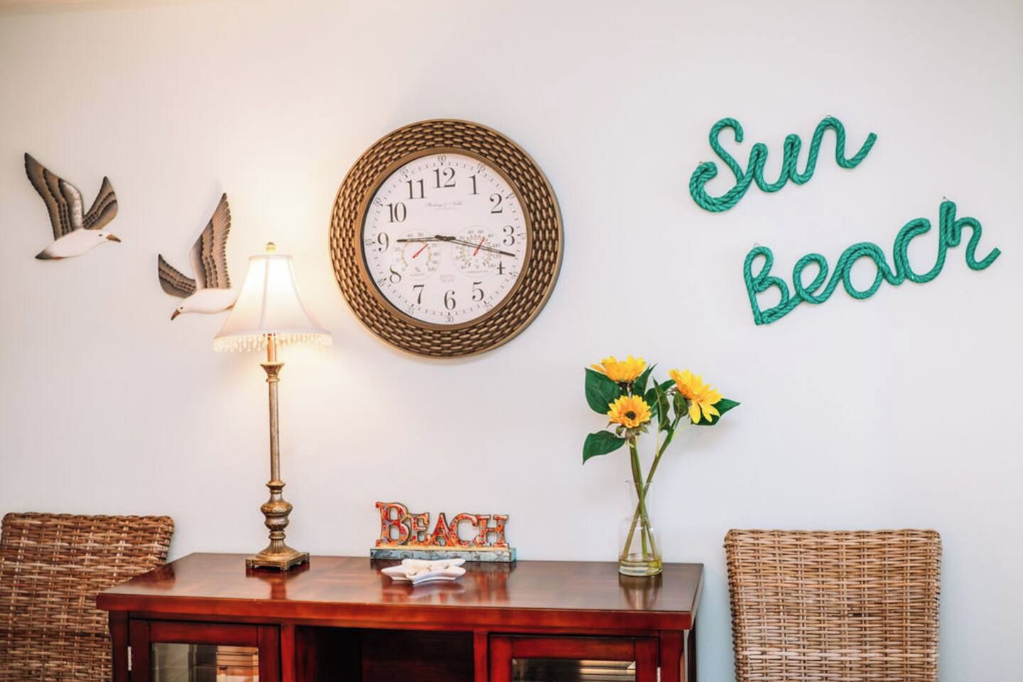 Una habitación con un reloj de pared, una lámpara, un jarrón con flores y un letrero que dice 'BEACH'.