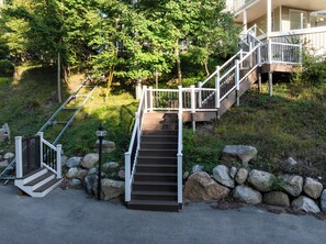 A set of outdoor wooden stairs leading to a building with a white railing, surrounded by greenery and rocks. - Beautiful Day | Large Grand Haven Retreat with Spectacular Lake Michigan Views + Beach (Grand Haven)