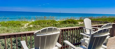 Una terraza con dos sillas Adirondack blancas que dan a una playa con dunas y el océano.