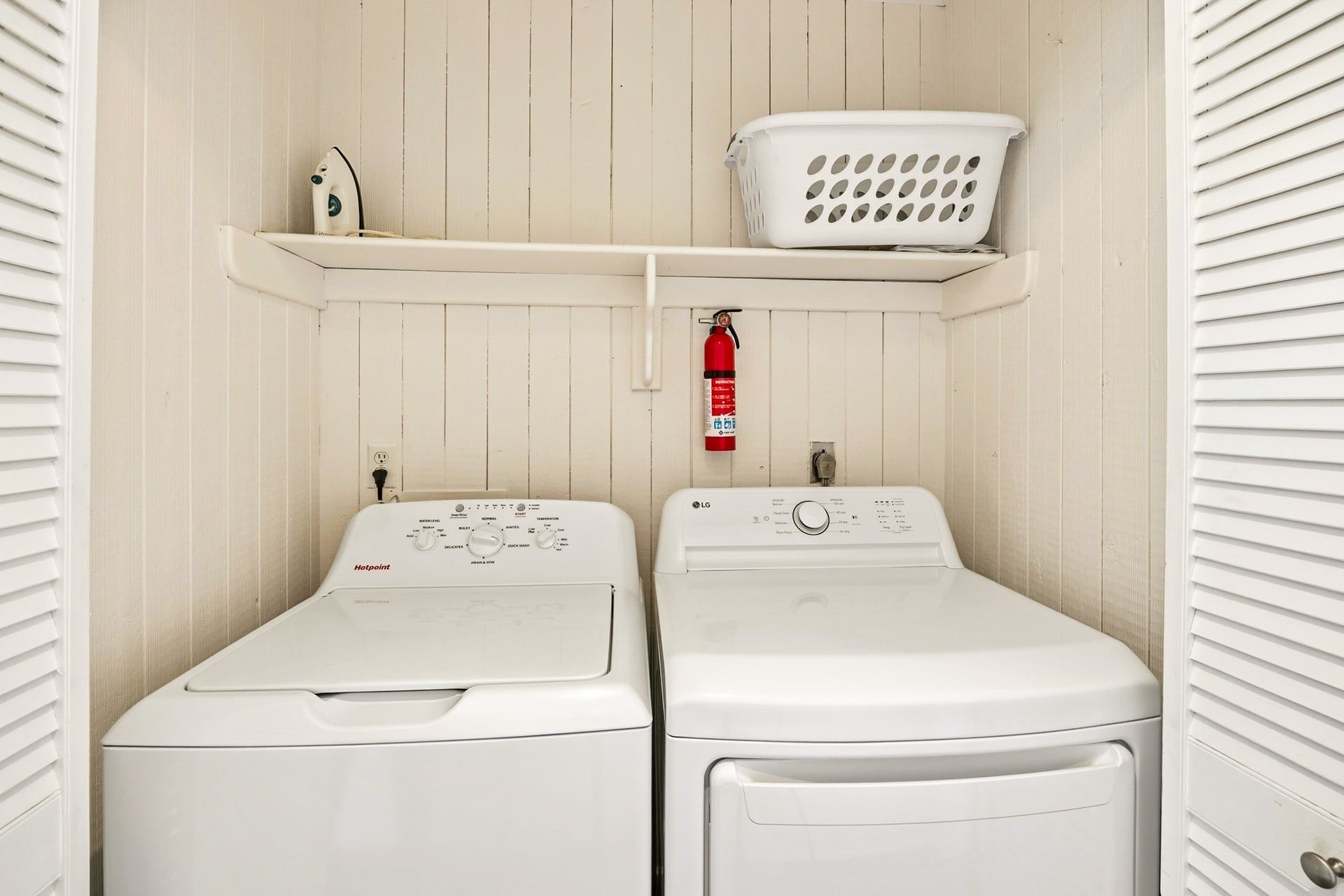 A laundry room with two front-loading washing machines, a white laundry basket on a shelf, a fire extinguisher, and a power outlet.