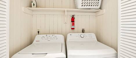 A laundry room with two front-loading washing machines, a white laundry basket on a shelf, a fire extinguisher, and a power outlet.